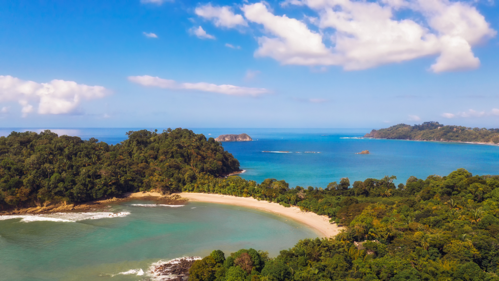 Aerial view of a curved sandy beach surrounded by dense green forest and turquoise waters—one of the best places to visit in Costa Rica, with scattered islands under a partly cloudy sky.
